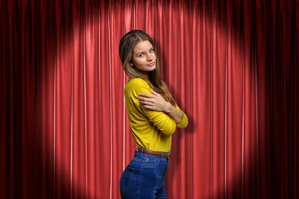 Young woman wearing blue jeans and yellow shirt embracing herself on red stage curtains background