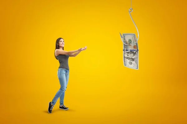 Jovem morena vestindo jeans casuais e t-shirt segurando suas mãos em dinheiro dólar no gancho de peixe no fundo amarelo — Fotografia de Stock