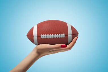 Açık mavi degrade arka planda Amerikan futbolu için büyük kahverengi top tutan ve bakan ve büyük kahverengi topu tutan kadın el Yan closeup.