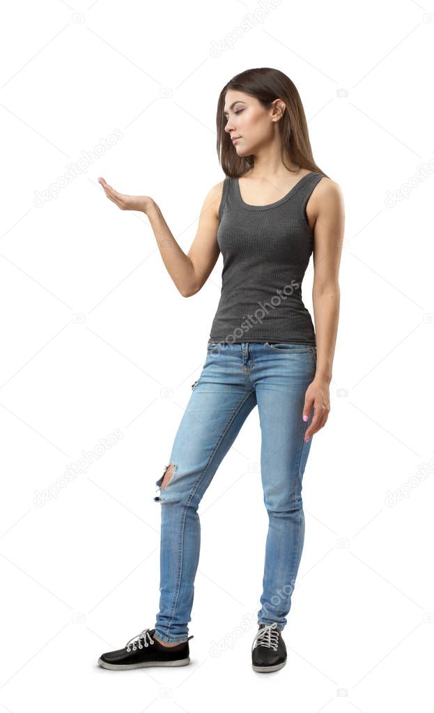 Young attractive woman in gray sleeveless top and blue jeans standing in half-turn with right arm bent at elbow and facing up isolated on white background.