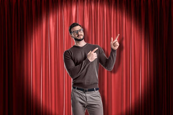 Front crop view of young handsome man in casual clothes standing in spotlight against red stage curtain and pointing up with both hands.