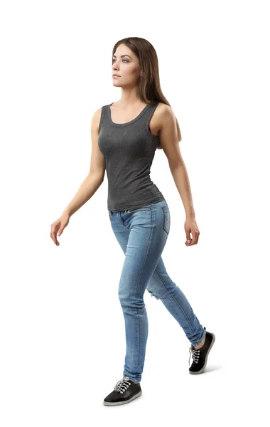 Junge attraktive Frau in grauem ärmellosen Top und blauen Jeans, die in halber Drehung geht und auf weißem Hintergrund isoliert zur Seite schaut. — Stockfoto