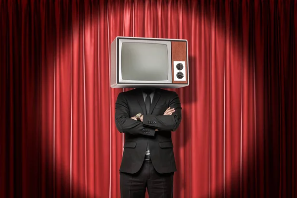 Front crop image of man in suit, with arms folded, and with TV set instead head, standing in spotlight at red stage curtain.