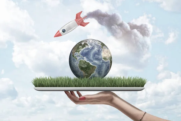Womans hand holding digital tablet with grass and planet Earth on screen, and rocket in air above leaving smoke trail behind. — Stock Photo, Image