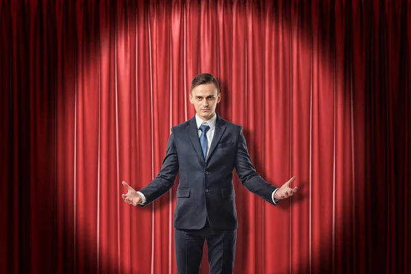 Young serious businessman with palms up on red stage curtains background