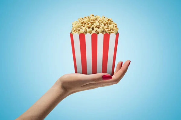 Seitliche Nahaufnahme einer Frauenhand, die nach oben zeigt und eine gestreifte Popcornbox auf hellblauem Hintergrund hält. — Stockfoto