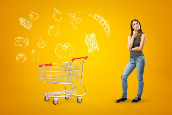 Young thinking brunette girl wearing casual jeans and t-shirt with shopping cart and cartoon food drawn on yellow background