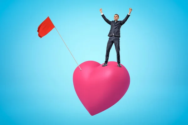 Jeune homme d'affaires heureux levant les bras sur grand coeur rose avec drapeau rouge sur fond bleu — Photo