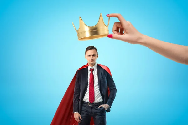 Kvinnlig hand Holding Golden Crown över ung affärsman med röd kappa på blå bakgrund — Stockfoto