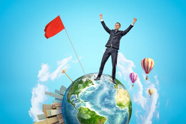 Small planet Earth with modern city popping up on one side and hot-air balloons flying in sky, and happy businessman who has planted red flag on planet