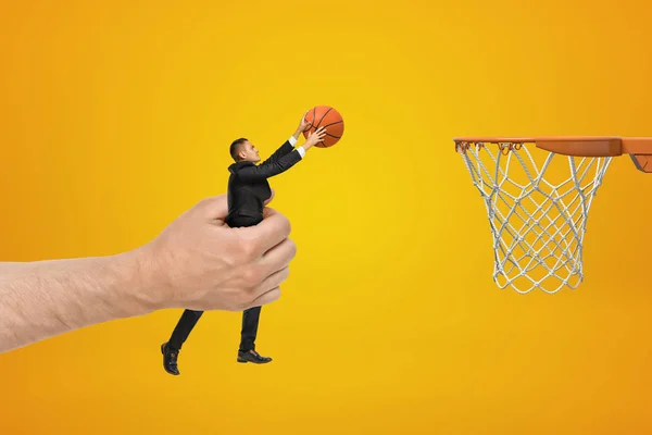 Grande mano masculina sosteniendo hombre de negocios con pelota de baloncesto llegar a aro sobre fondo amarillo — Foto de Stock