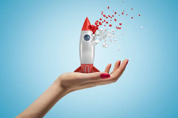 Kant close-up weergave van dames hand naar boven, holding Toy Rocket die begint te disslove in stukken, op licht blauwe achtergrond. — Stockfoto