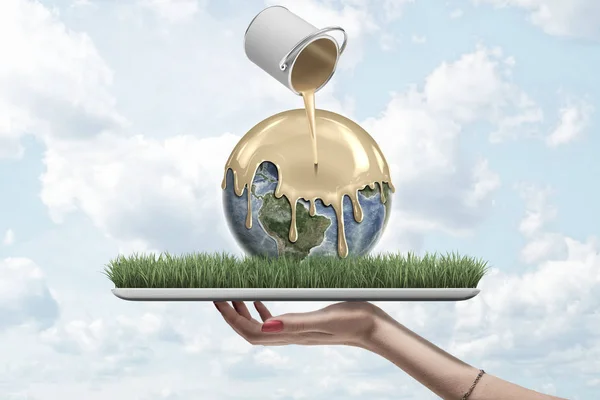 Closeup of womans hand facing up holding digital tablet with Earth lying on screen and paint can in air pouring gold paint onto Earth.