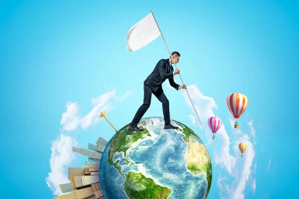 Small planet Earth with modern city popping up on one side and hot-air balloons flying in sky, and businessman walking on planet and carrying white flag.