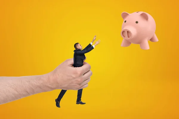 Grote hand met kleine zakenman reiken met zijn beide handen voor schattig roze Piggy Bank drijvend in lucht op Amber achtergrond. — Stockfoto