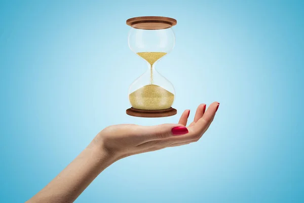Side closeup of womans hand facing up and levitating hourglass on light blue background. — Stock Photo, Image