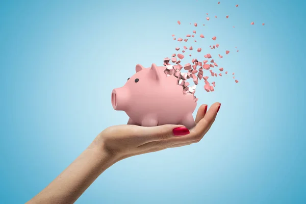 Close-up of womans hand facing up and holding cute pink piggy bank that has started to disintegrate into pieces on light-blue background.