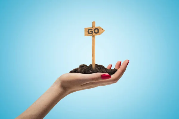 Close-up of womans hand holding palmful of soil with small wooden road sign that reads GO stuck into it on light-blue background. — Stock Photo, Image