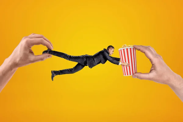 Big male hand holding tiny businessman who is reaching to another big hand holding popcorn bucket on yellow background