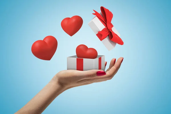 Πλευρά κοντινό πλάνο των γυναίκες χέρι μπροστά και κρατώντας ένα μικρό λευκό κουτί δώρου, το καπάκι του στον αέρα και χαριτωμένες κόκκινες καρδιές που πετούν στο γαλάζιο φόντο. — Φωτογραφία Αρχείου