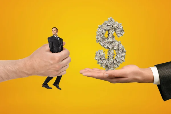 Велика чоловіча рука передає крихітного бізнесмена в іншу руку з великими грошима знак долара на жовтому тлі — стокове фото