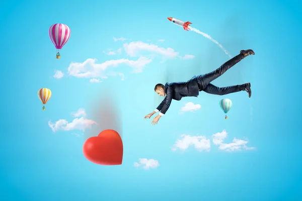 Businessman reaching to big red heart with hot air balloons and silver red space rocket in the air on blue background