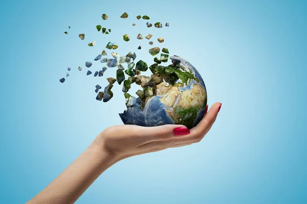 Closeup of womans hand holding Earth which is breaking into small pieces that are flying away on light blue background. — Stock Photo, Image