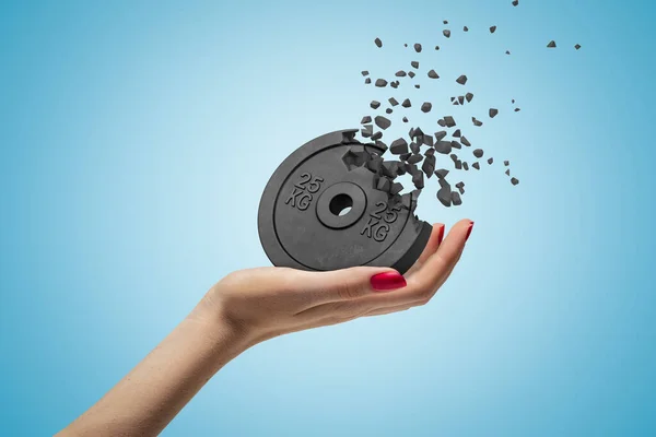 Side closeup of womans hand facing up and holding black 25kg weight plate that is dissolving in pieces on light blue background. — Stock Photo, Image