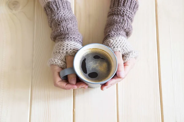Woman hands with wool warmers holding a cup of coffee on wooden background, top view