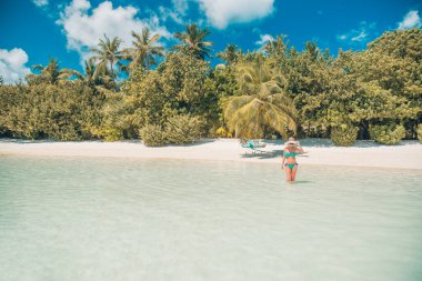 Cennet adasında lüks tatil beldesi. Mavi denizin üzerindeki palmiye ağaçları, egzotik varış yerleri, tatil yerleri. Plaj manzarası, kadın hasır şapka ve mavi yeşil bikini seksi bacaklar. Güzel kadın turist yaz tatili tatilinde dinleniyor.