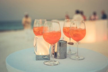 Plaj partisinde aperol spreyli kokteyller ve arka planda bulanık insanlar vardı. Sosyal etkinlik kaprisli afiş tasarımı sığ odak noktasında. Plaj partisinde bulanık insanlarla plaj günbatımı manzarası mum ışığında beyaz masadaki aperol içeceklerine odaklan