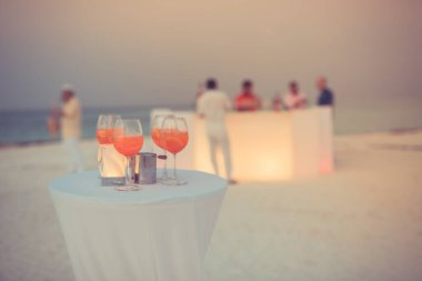 Plaj partisinde aperol spreyli kokteyller ve arka planda bulanık insanlar vardı. Sosyal etkinlik kaprisli afiş tasarımı sığ odak noktasında. Plaj partisinde bulanık insanlarla plaj günbatımı manzarası mum ışığında beyaz masadaki aperol içeceklerine odaklan