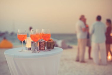 Plaj partisinde aperol spreyli kokteyller ve arka planda bulanık insanlar vardı. Sosyal etkinlik kaprisli afiş tasarımı sığ odak noktasında. Plaj partisinde bulanık insanlarla plaj günbatımı manzarası mum ışığında beyaz masadaki aperol içeceklerine odaklan