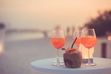 Plaj partisinde aperol spreyli kokteyller ve arka planda bulanık insanlar vardı. Sosyal etkinlik kaprisli afiş tasarımı sığ odak noktasında. Plaj partisinde bulanık insanlarla plaj günbatımı manzarası mum ışığında beyaz masadaki aperol içeceklerine odaklan