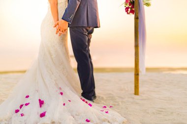 Gelin ve damat gün batımı kumsal manzarasında, çiçekler beyaz elbisede. Gün batımında romantik bir sahil düğünü. Lüks düğün ve balayı