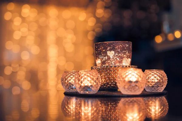 Beautiful candle light with table reflection and golden blur bokeh lights background. Christmas holiday cozy home interior closeup. Romantic glass jars with burning candles on the dark background