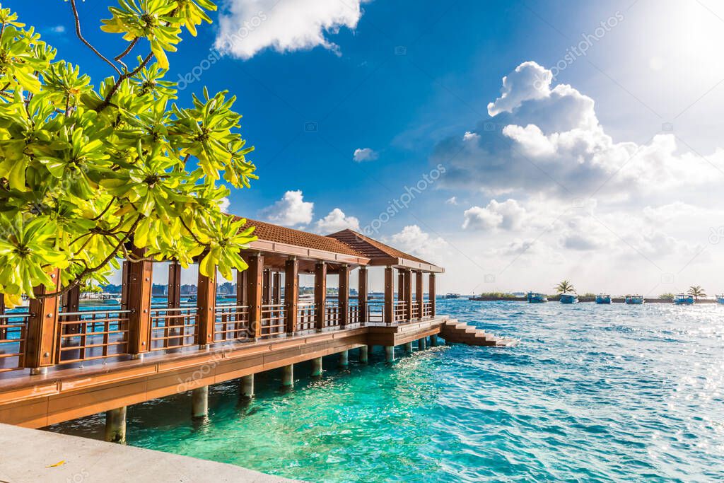 Perspective view of a wooden pier on the tropical seashore with clear blue sky with some white clouds and sea with turquoise water. Maldives capital, Male city, luxury dock with jetty for transportation, speedboats and yachts