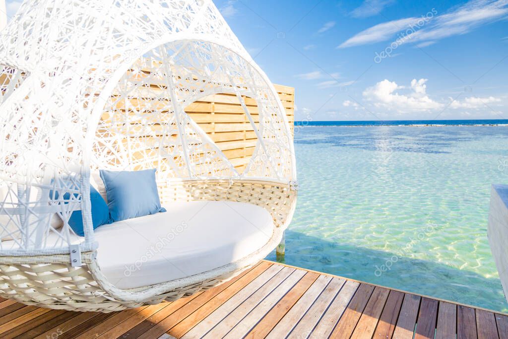 Tropical beach background as summer landscape with beach swing over calm sea, view from over water villa and wooden deck. White luxury lounge swing in Maldives shoreline. Exotic travel destination, fun and love couple vacation or travel background