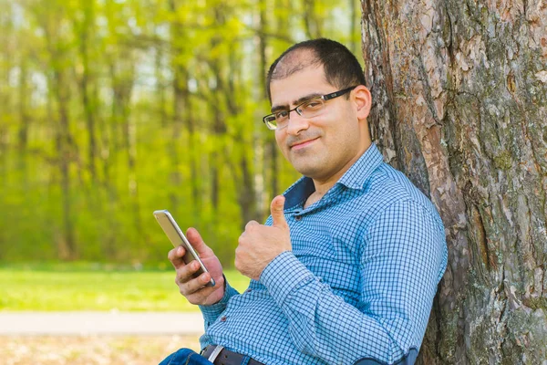 bir telefonla onun elinde bir ağacın altında oturup ve gülümseyen bir adam