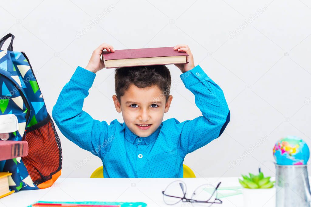 A dark haired little boy in a blue shirt sits at a table and holds a book on his head. Attention deficit in preschool children..