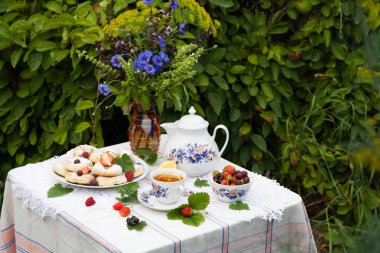 Bahçede masada çiçek, çay, kurabiye ve çilek buketi ile natürmort