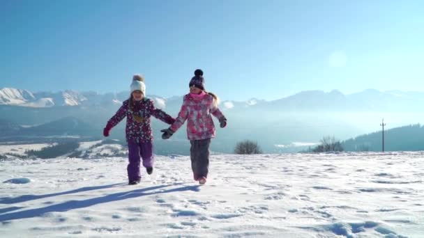 Очаровательные дети в наружной одежде бегают по заснеженной местности с красивыми горами на заднем плане под солнцем — стоковое видео