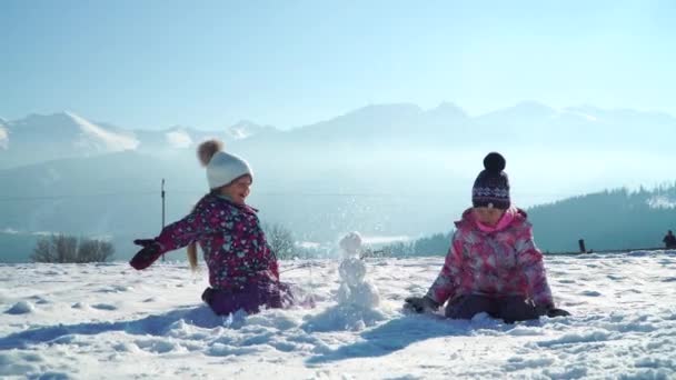 背景に山を持つ日光の雪のフィールドで遊んでいる間、outwear の子供たちは小さな雪だるまを作る — ストック動画