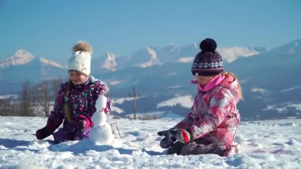 Barn i ytterkläder gör små snögubbe medan du spelar på snöiga fält i solljus med berg på bakgrunden — Stockvideo