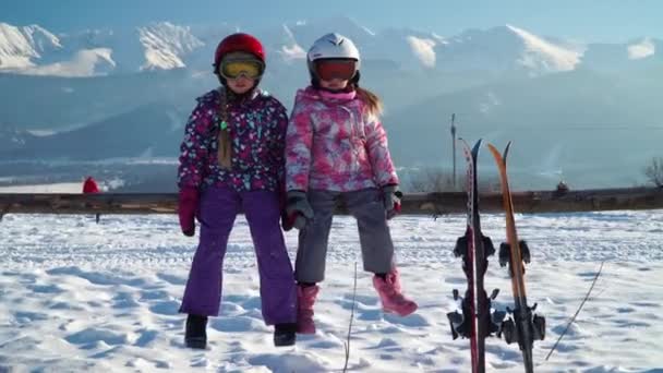 山に対してスキー近くの木製フェンスに座ってヘルメットで outwear とマスクの小さな女の子 — ストック動画