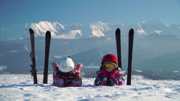 Kinderen in helmen en brillen liggend op besneeuwde terrein met ski's in de buurt op de achtergrond van de bergen — Stockvideo