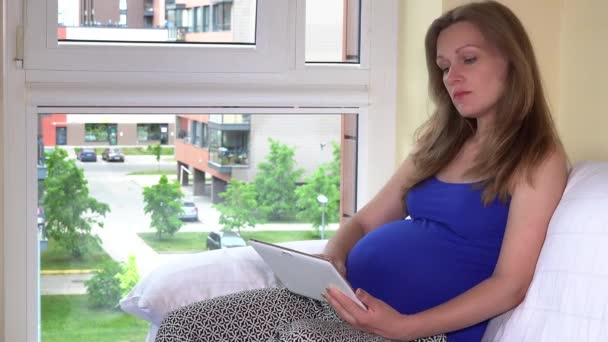 Gravid kvinna i blå skjorta använder surfplatta på hem sitter nära fönstret — Stockvideo