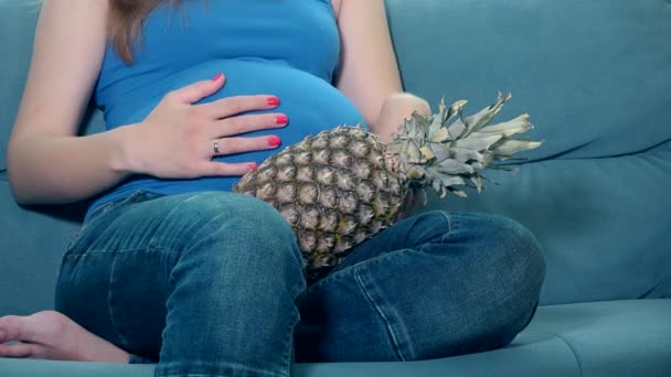 Těhotná žena tahu velké bříško a podržte ananas ananas plody sedí na pohovce — Stock video