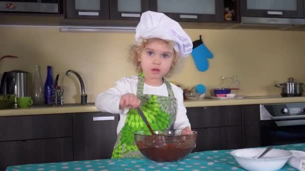 Criança menina grave com colher misturando massa em tigela de vidro na mesa da cozinha — Vídeo de Stock