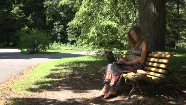 学生の女の子は、緑豊かな公園のベンチの上に座ってのラップトップで動作します。4 k — ストック動画